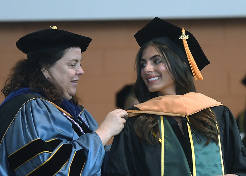 Nancy George on the left wearing a blue and black graduation regalia gown placing a gold hood on a woman on the right wearing a green stole with gold embroidery, gold tassel, and a black cap and gown.