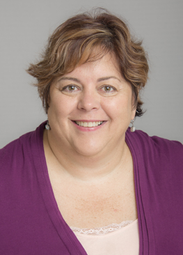  Wayne State University College of Nursing Associate Professor Clinical Nancy George smiles in a head shot photo wearing a purple shawl on her shoulders, peach shirt, silver earrings and auburn short hair in front of a gray wall.