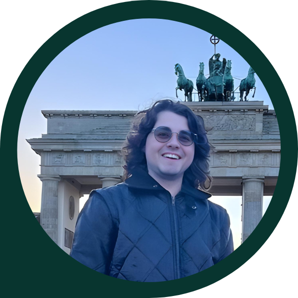 Man standing in front of the Brandenburger Tor in Berlin, smiling wearing sunglasses, a leather jacket and jeans.