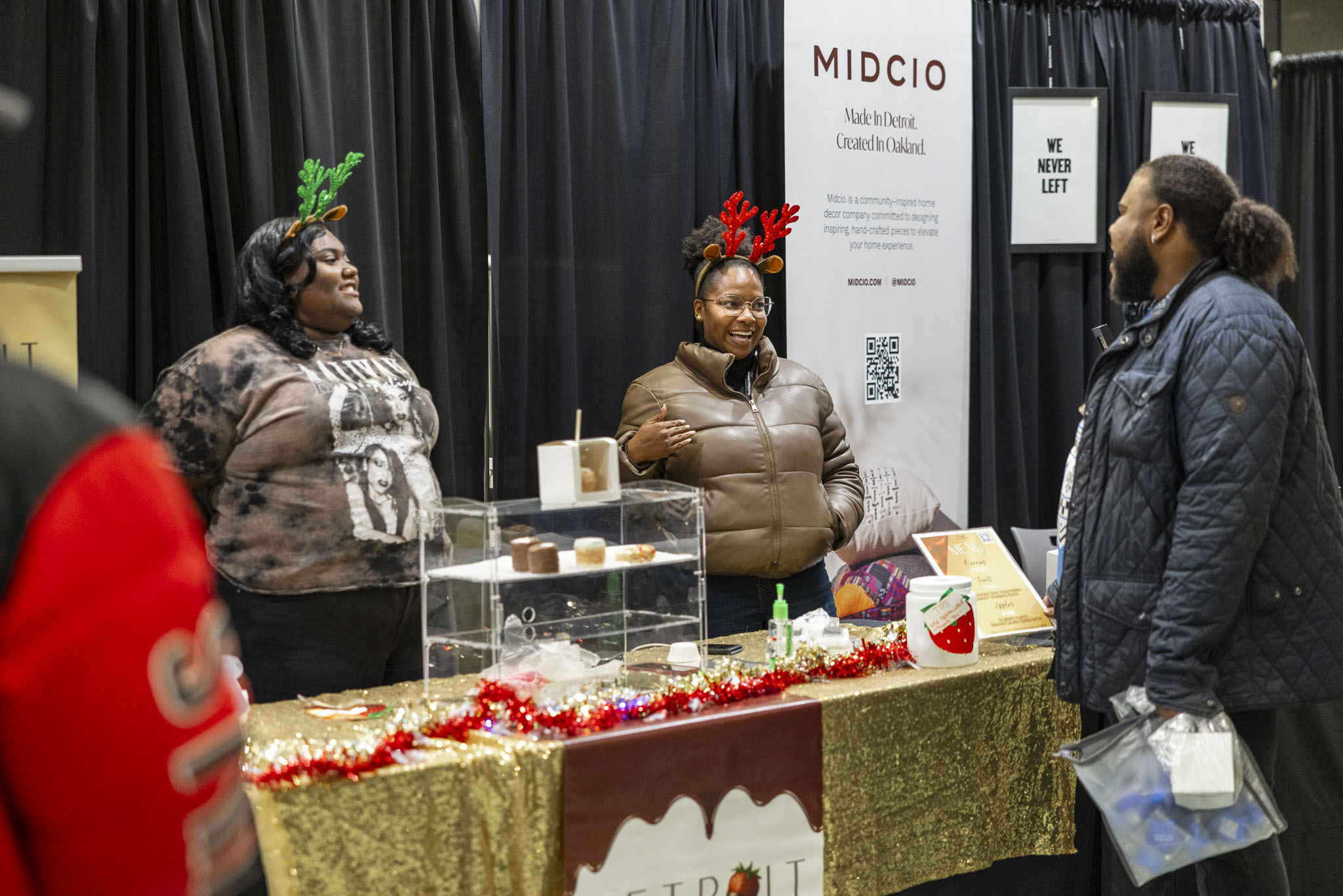 Three people interact around a vendor table display for the business Detroit Berry Company. The table features an array of desserts