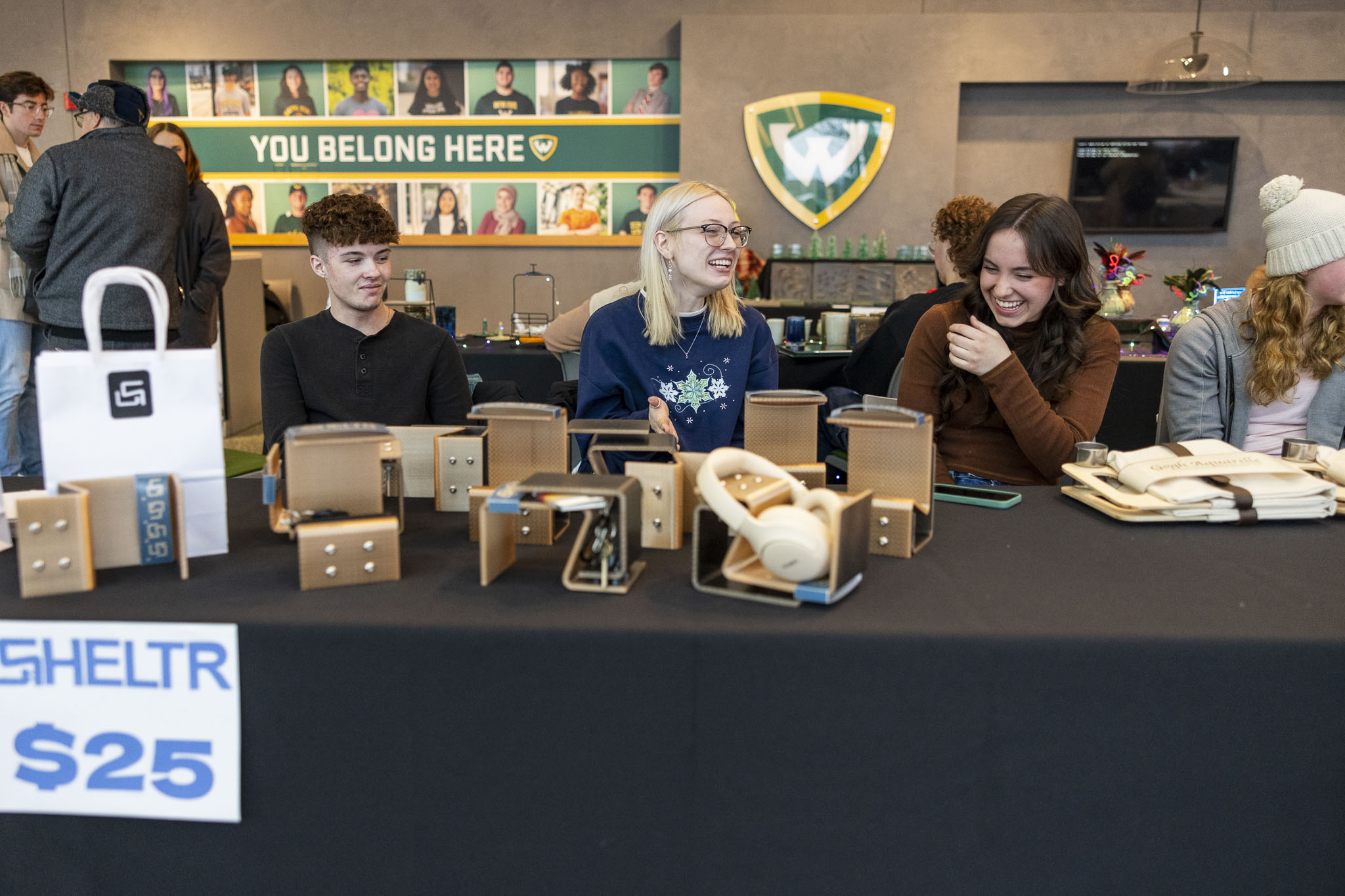 Three people talk amongst each other while sitting behind a vendor table display for their project SHELTR