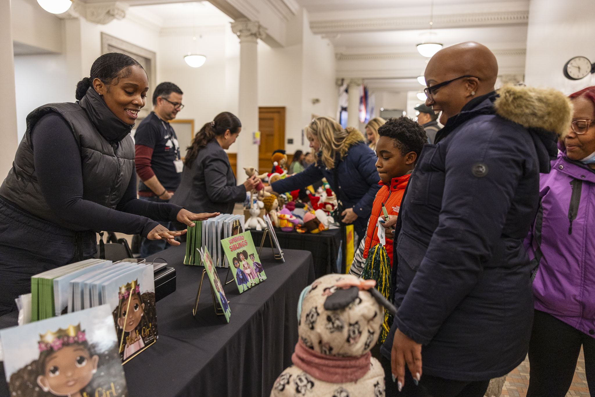 A person shows children's books from their vendor table display to an adult and two children