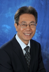 A headshot of Zhang. He is pictured wearing glasses and a dark suit and tie in front of a blue background. 